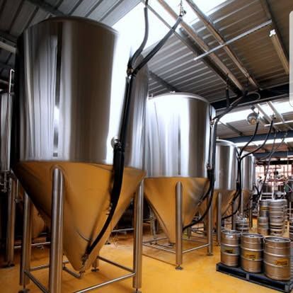 Shiny metal brewing tanks in a brewery with beer kegs and piping.