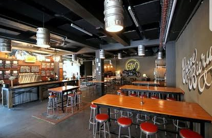 Interior of a bar with industrial decor. Wooden tables, red stools, keg-shaped lights, and a dark ceiling.