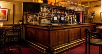 A wooden bar with many liquor bottles, taps, and bar stools. Red carpet, warm lighting.