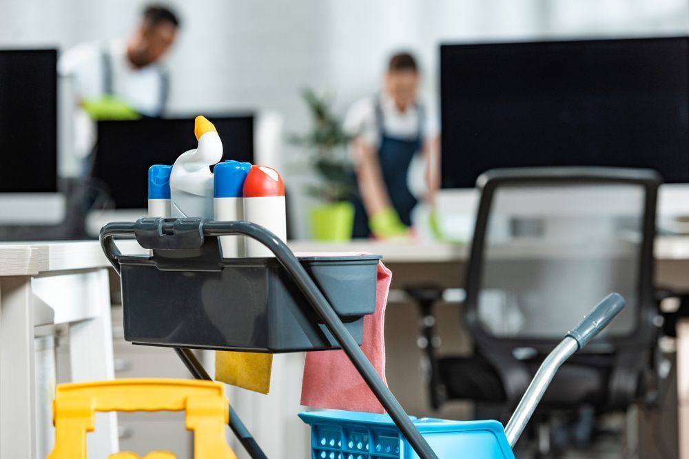 Cleaning crew in office; cleaning cart with supplies in foreground, workers in background.