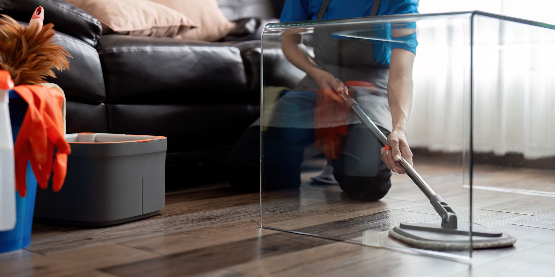 Person mopping a wooden floor. There is a bucket with cleaning supplies. Black leather sofa in the background.