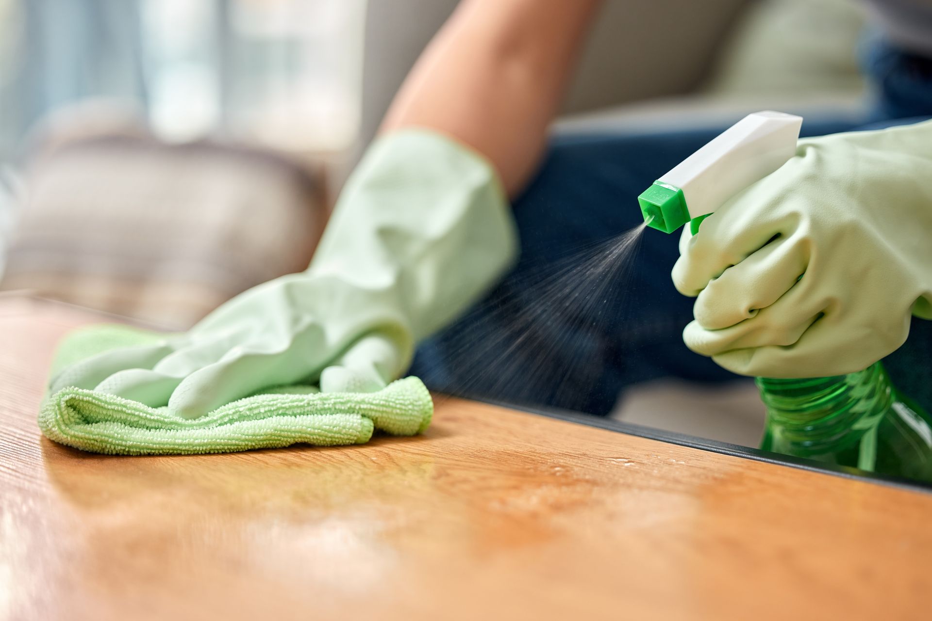 Person wearing green gloves spraying cleaner onto a wooden surface and wiping it with a green cloth.