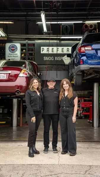 Three people posing in a car garage with lifted cars and a “PREFER” sign in the background.