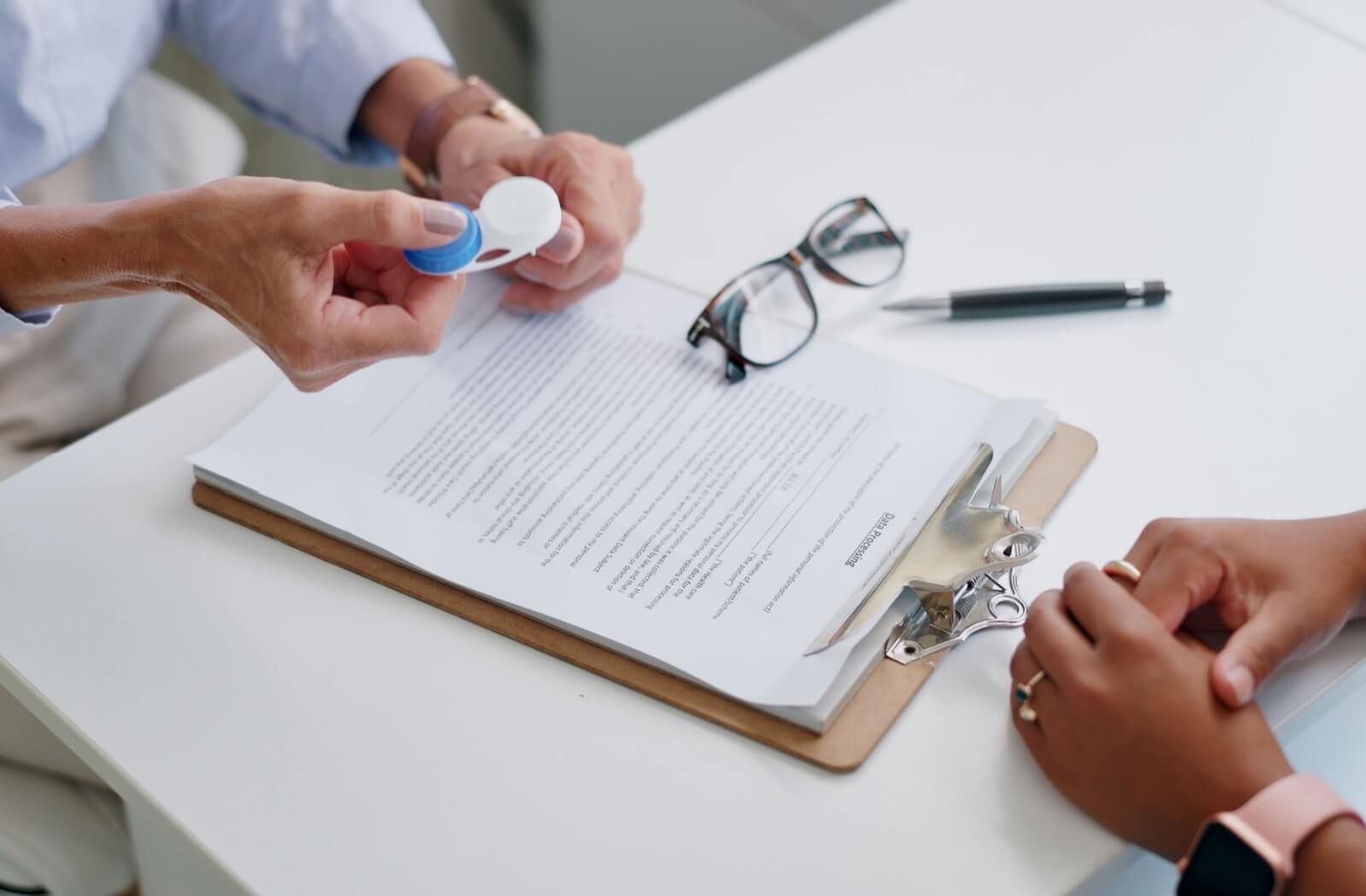 Image of a pair of hands holding a pair of contact lenses and another pair of hands folded near note pad