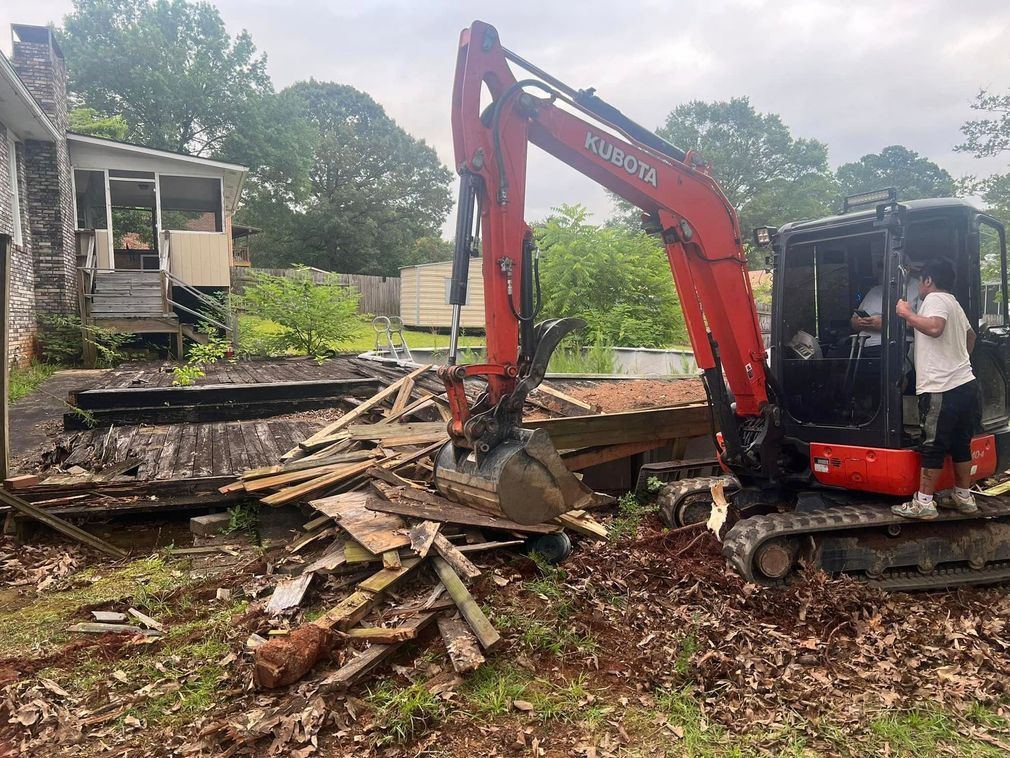 Excavator clearing debris after demolition beside a damaged house in a muddy yard