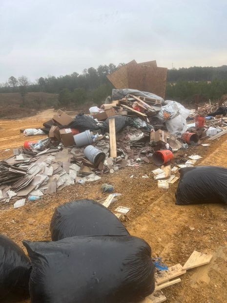 Dumped trash pile in a dirt lot, with black bags and scattered debris near trees and a brick wall.