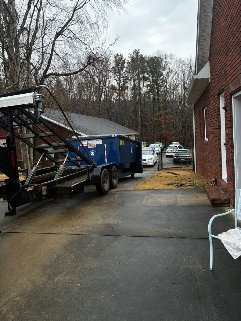 Blue dumpster trailer in a wet parking lot beside a brick building, with cars and trees in the background