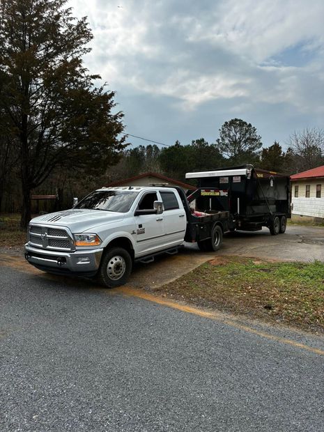 White work truck hitched to a black trailer on a gravel driveway near trees and a house.