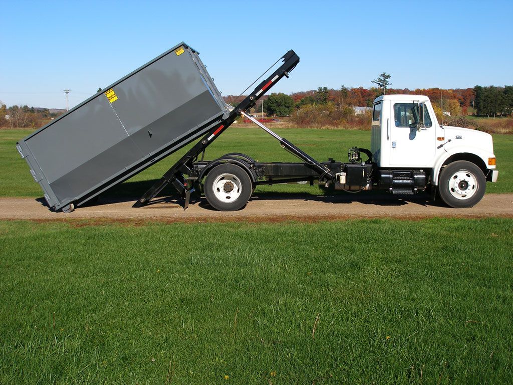 White tow truck tipping a large gray dumpster in a grassy field under a clear sky