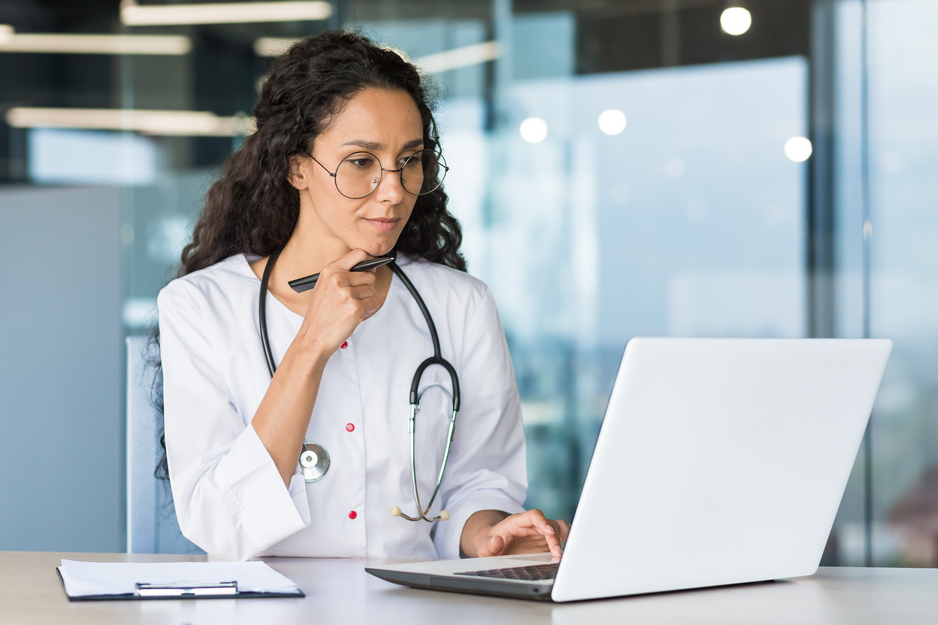 A healthcare worker with long curly hair resting her chin on her hand while looking at her laptop intently.