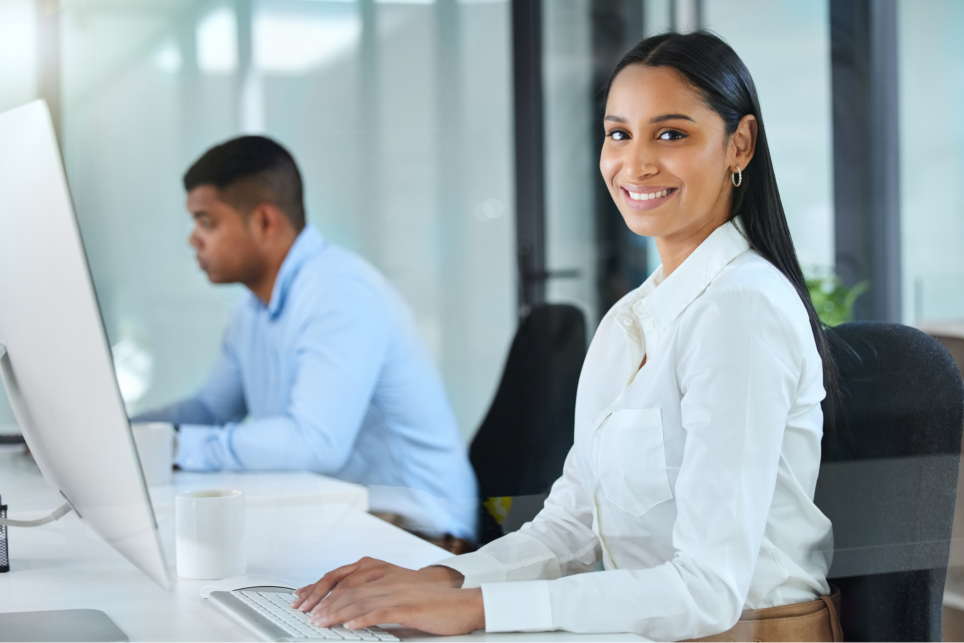 A woman in a corporate attire sitting straight and smiling while typing on a keyboard.