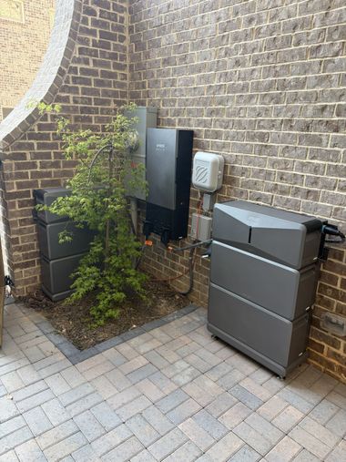 Brick corner with utility boxes, a gray bin, and a small leafy plant on paved ground.