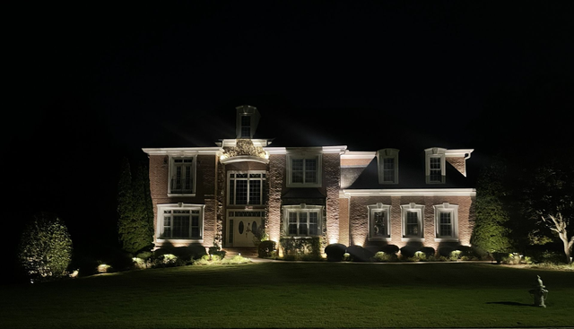 Large brick house lit at night, with glowing windows and a manicured lawn.