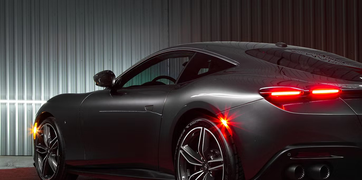 Dark gray sports car, lit by headlights, parked in front of a silver metal wall.