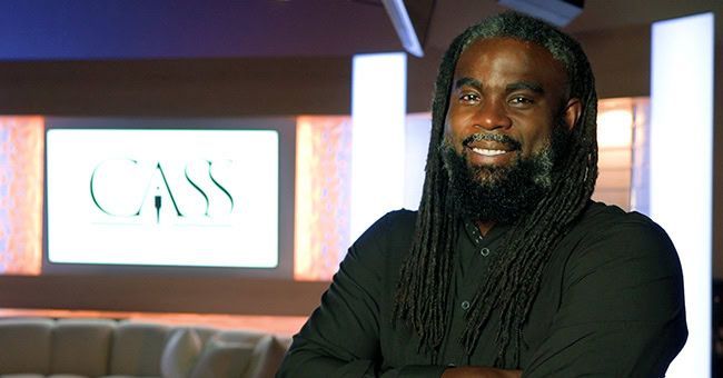 Man with dreadlocks and beard smiles, standing in a lounge-like setting with the word