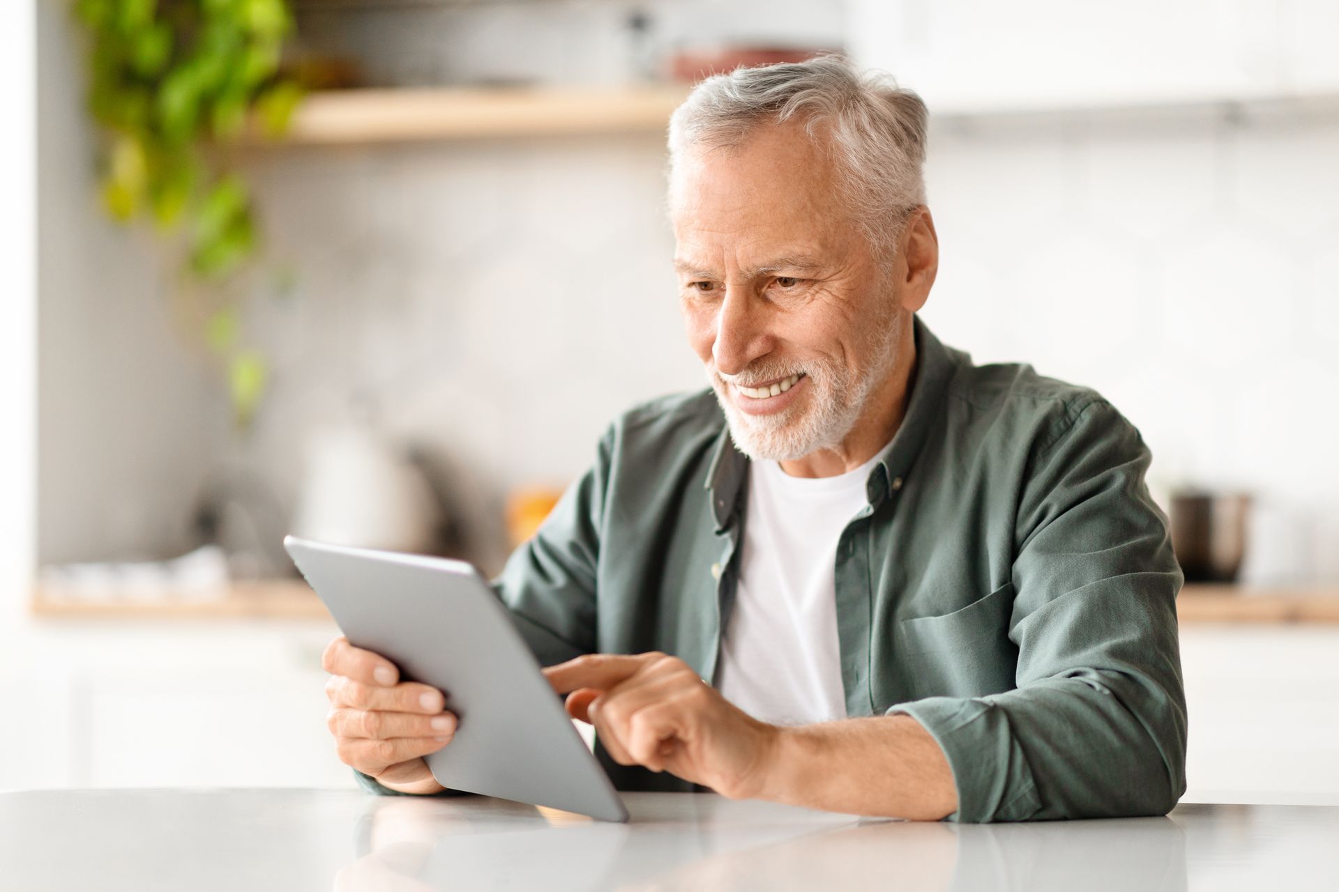 An older man with gray hair smiles while using a tablet at a kitchen counter.