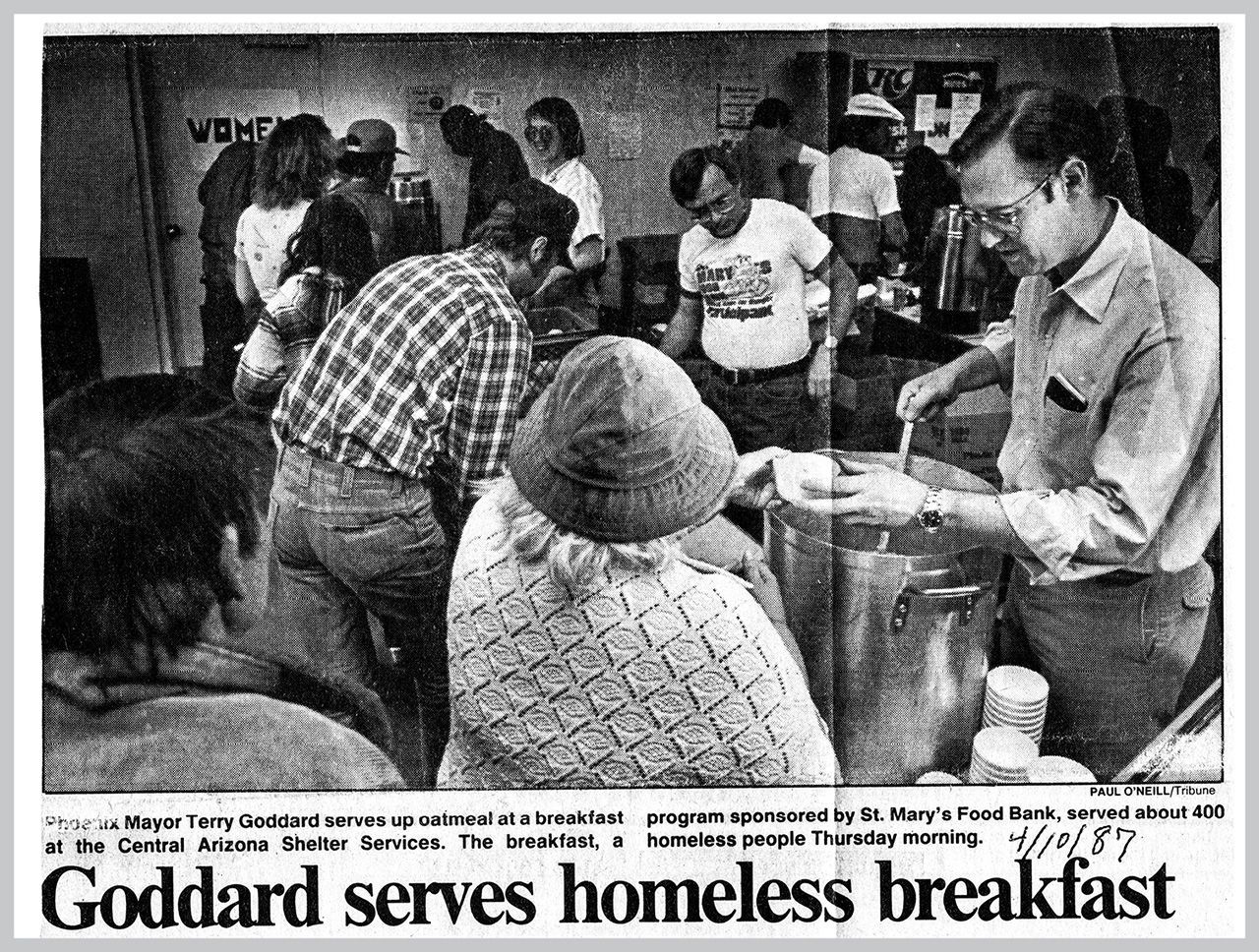 Men serve breakfast at a food bank. People line up; a man in glasses serves soup.
