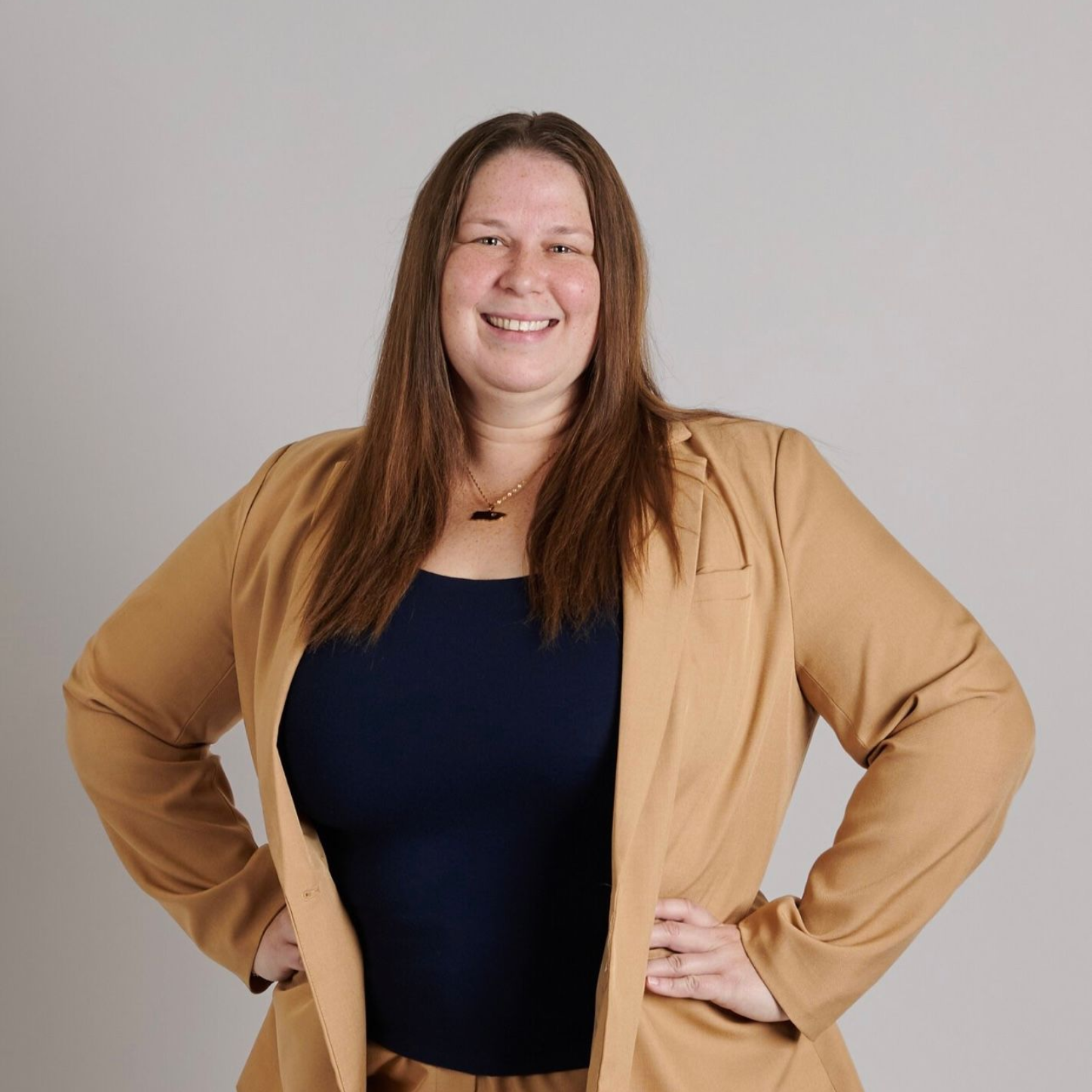 Woman in tan blazer and navy top, smiling with hands on hips.