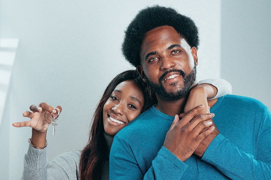 Couple smiles, holding keys in new home, woman's arm around man's neck, light background.