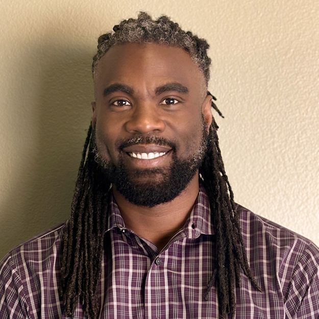 Man with dreadlocks and beard smiles, wearing a purple plaid shirt.