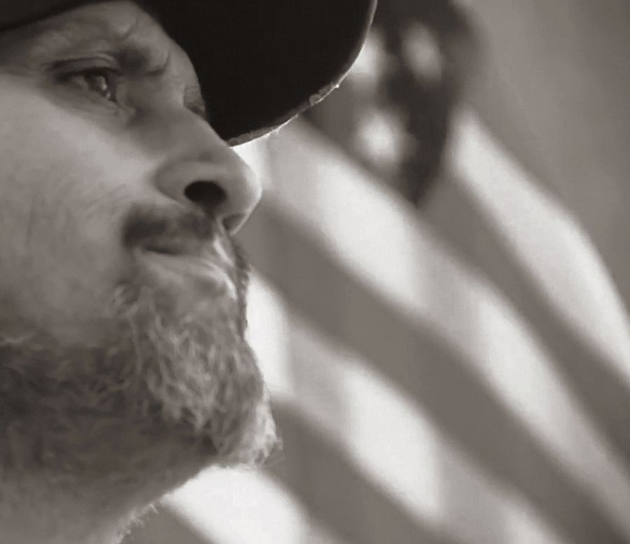 Man with a beard and hat looking leftward, American flag blurred in the background.