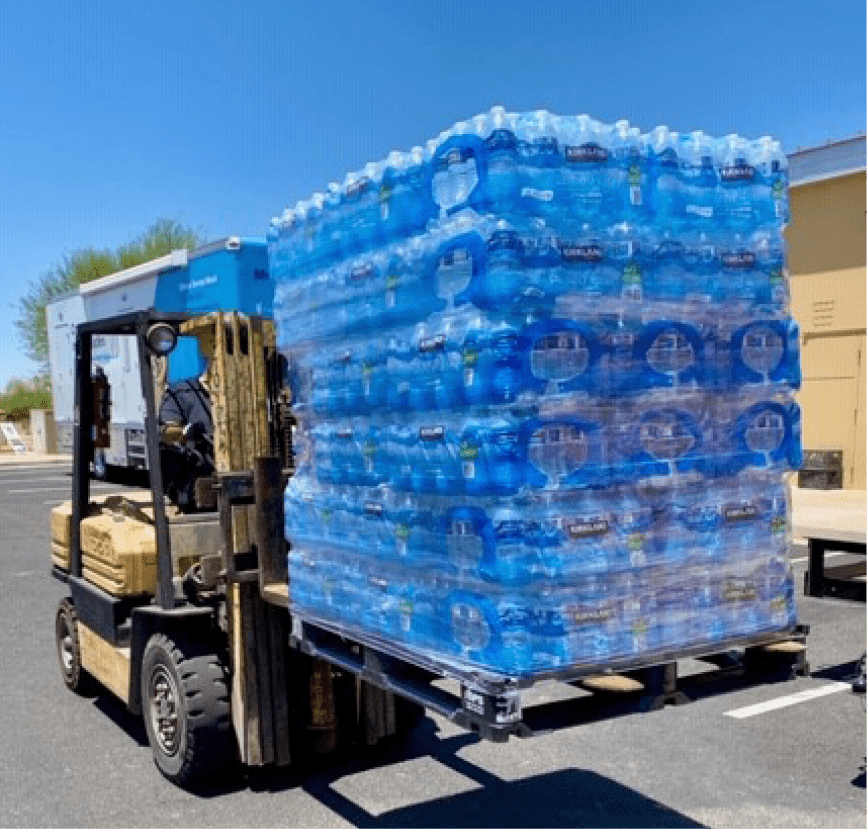 Forklift carrying a pallet stacked high with bottled water. Outdoors, sunny.