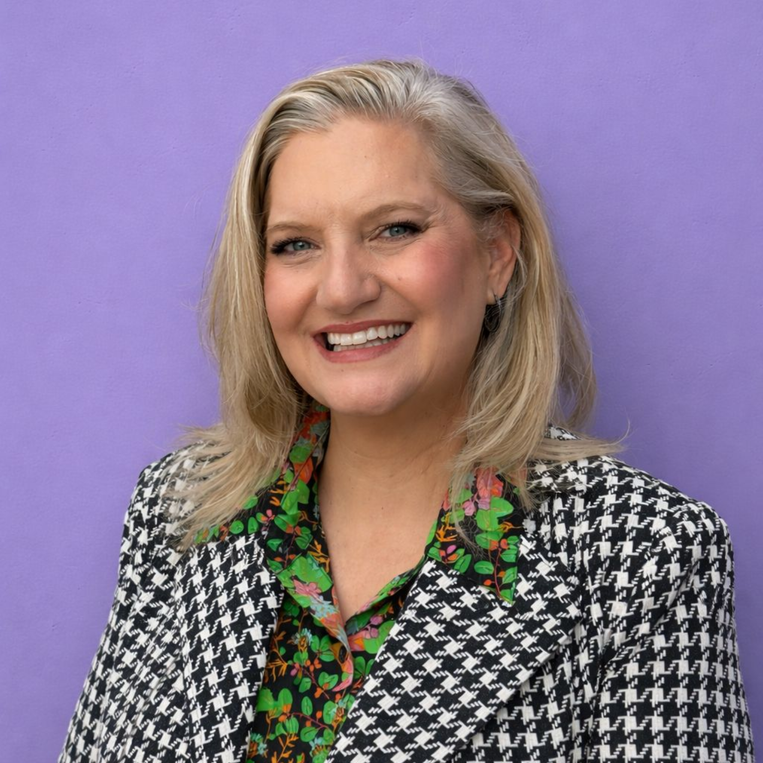 Woman smiling, wearing a black and white houndstooth jacket and floral top, against a purple background.