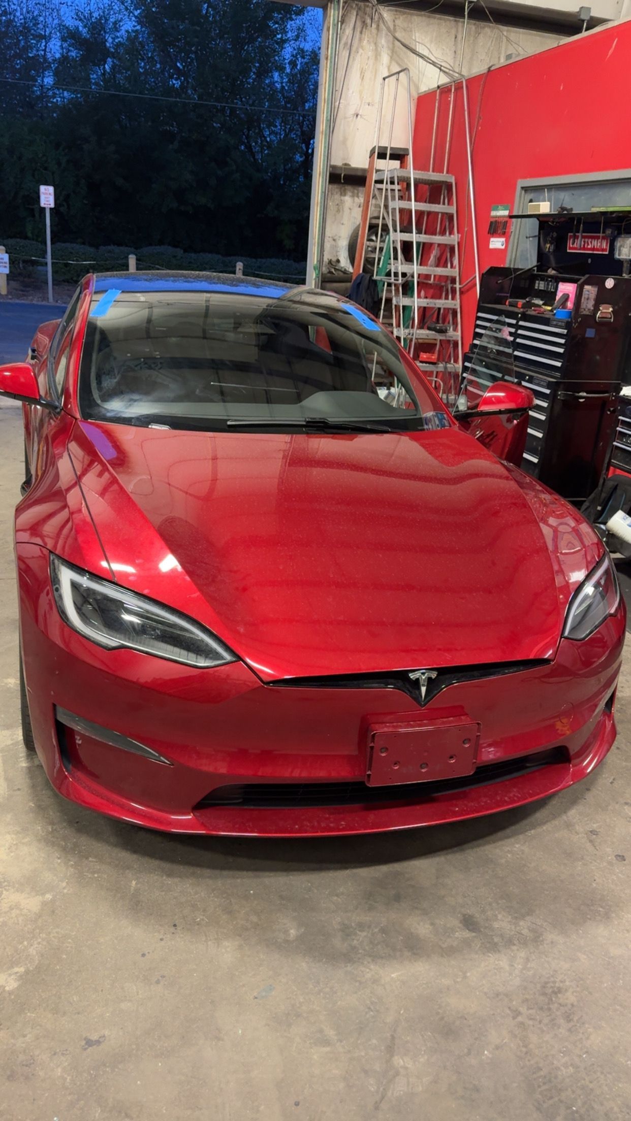 Red Tesla Model 3 parked indoors in a garage, front view with its hood open slightly visible.