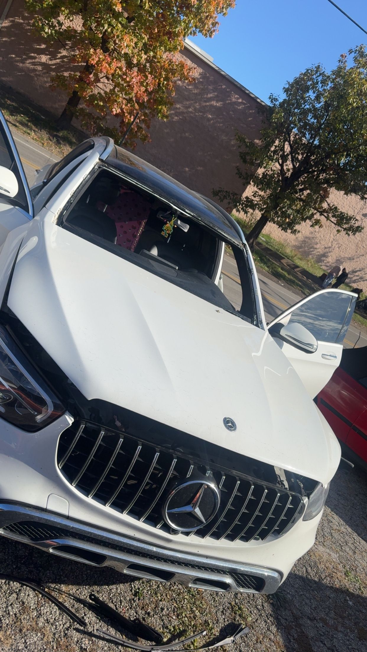 White Mercedes SUV with both front doors open, parked outdoors near trees and a brick building.
