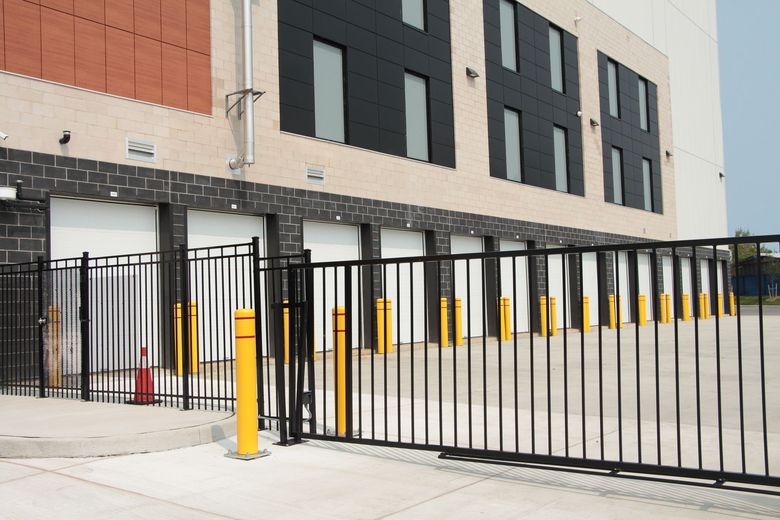 A row of white loading dock doors at a modern facility behind a black metal security fence and yellow bollards.