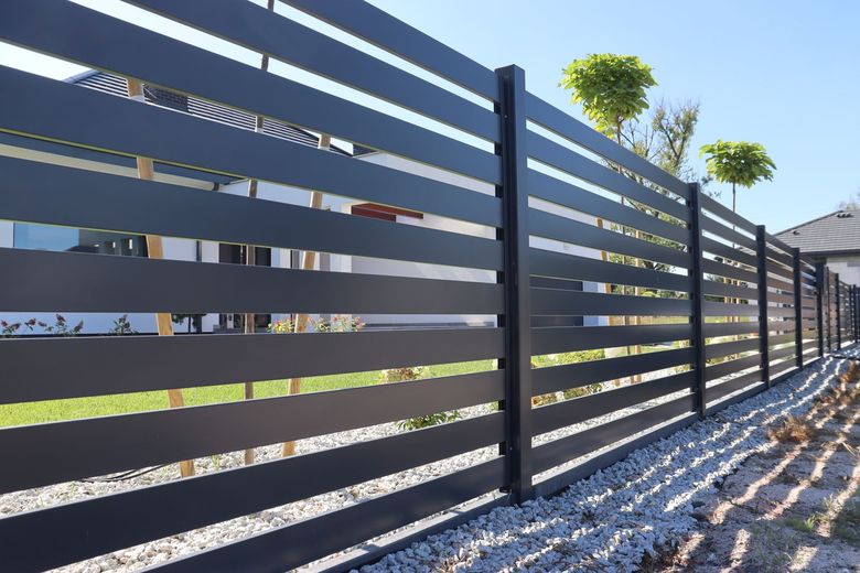 A modern black horizontal slat fence lines a residential property with a green lawn and gravel ground.