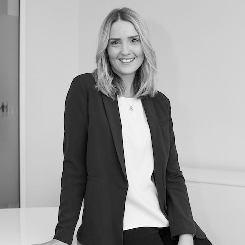 Smiling woman in a black blazer and white top leaning against a white desk indoors
