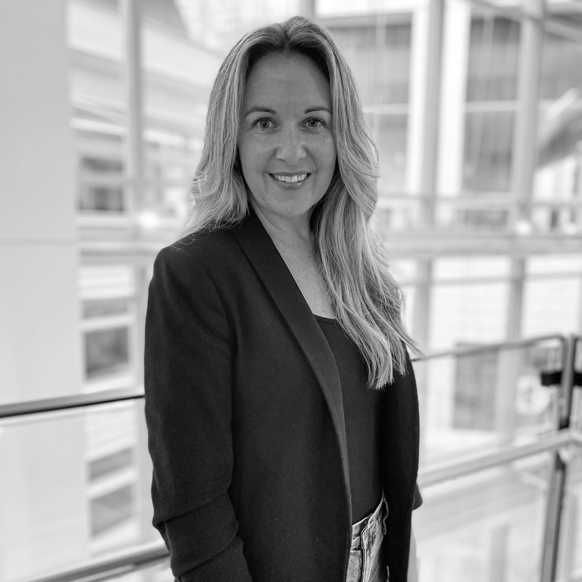 Smiling woman in a blazer standing in a bright office hallway with glass walls