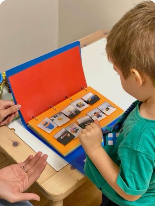 Un adulto ayuda a un niño a leer un libro naranja en una mesa con útiles escolares.