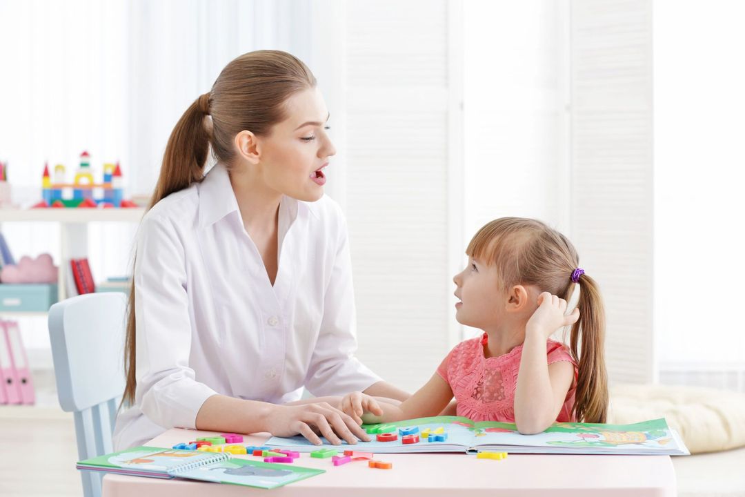 Una maestra y un niño conversan en una mesa de aula con materiales didácticos coloridos.