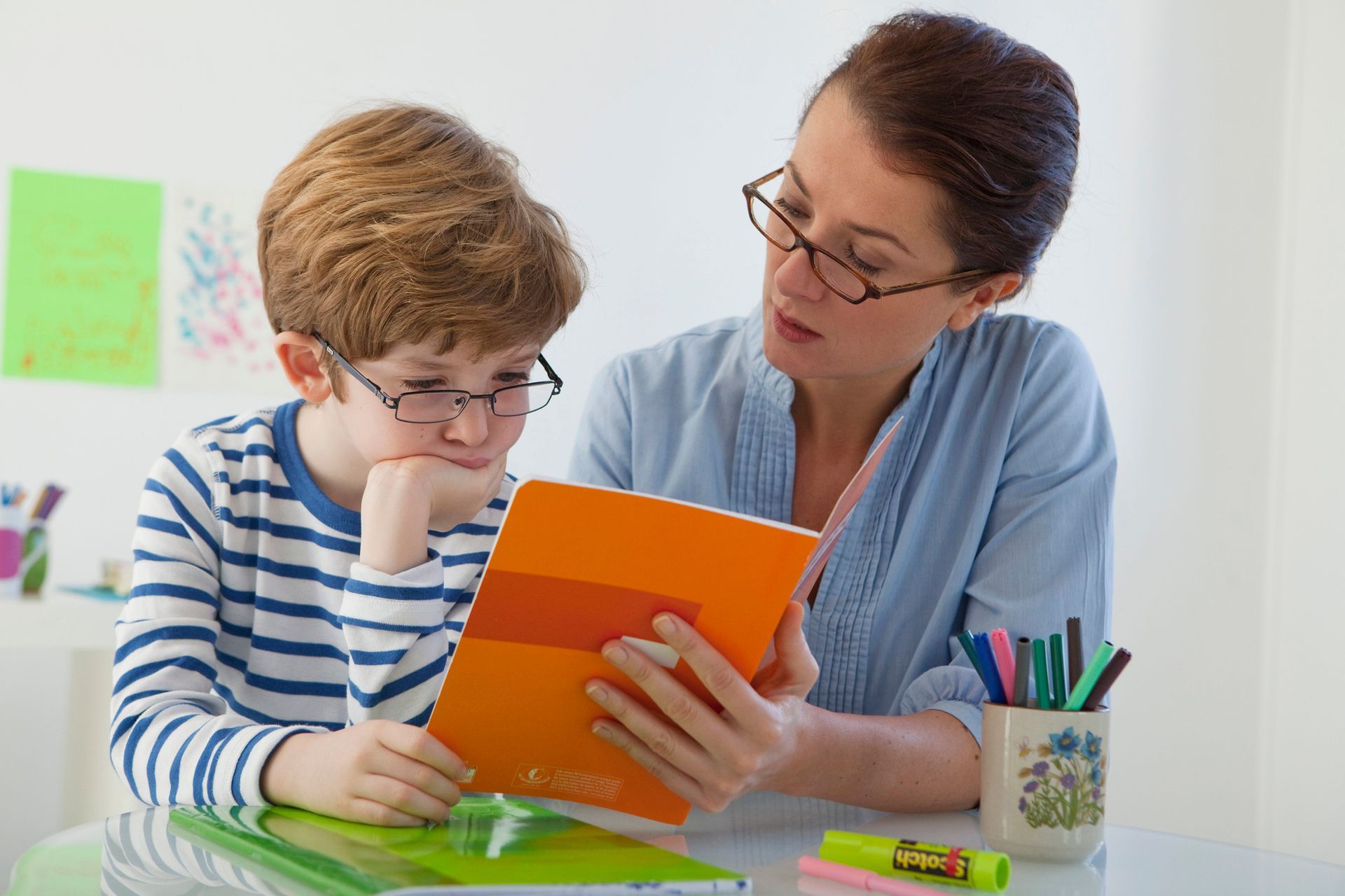 Un adulto y un niño leen un libro naranja en una mesa con material escolar.