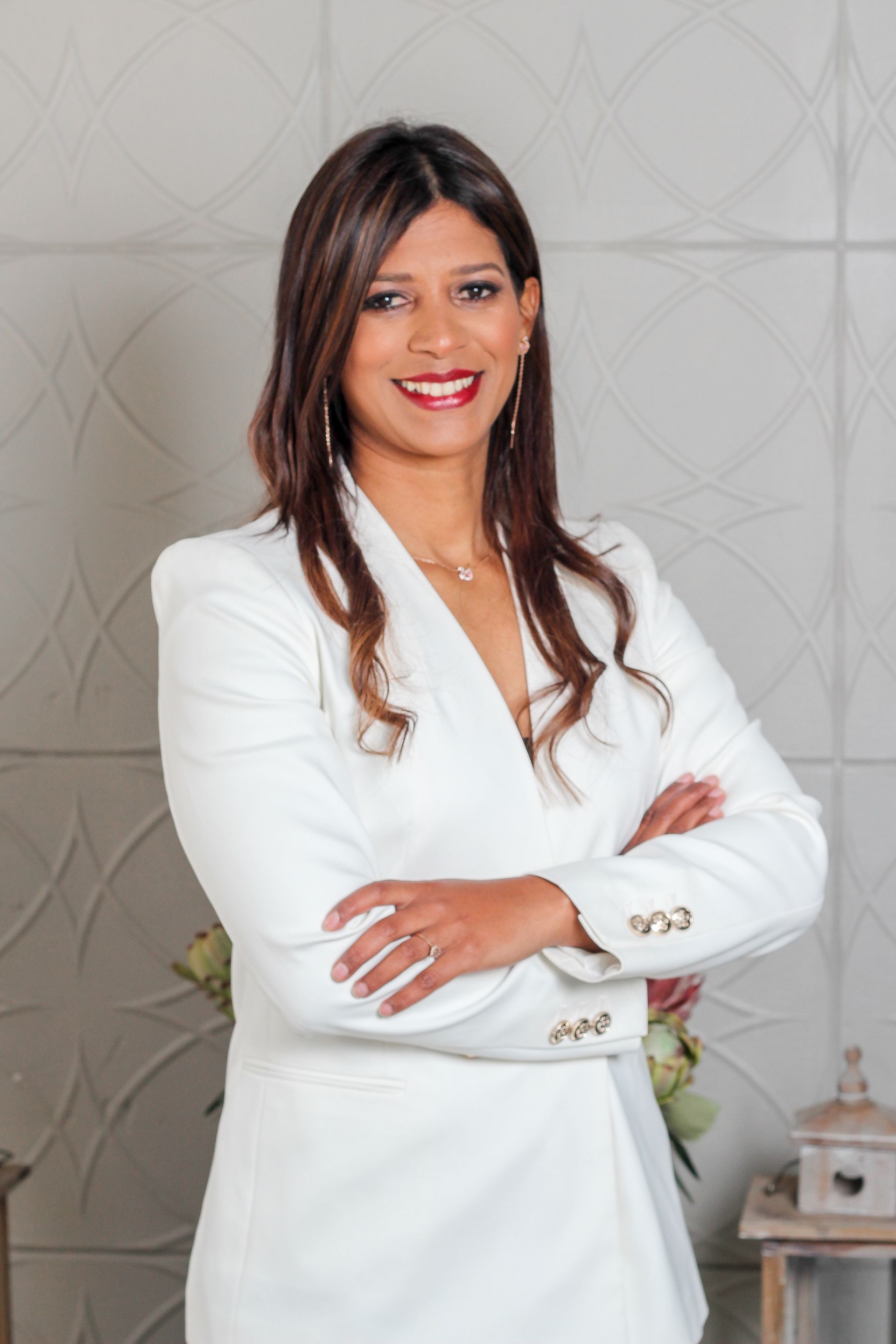 Woman in a white suit smiling with arms crossed against a light patterned wall.