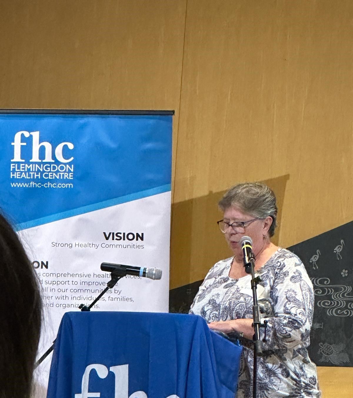 A woman is speaking into a microphone in front of a banner that says fhc