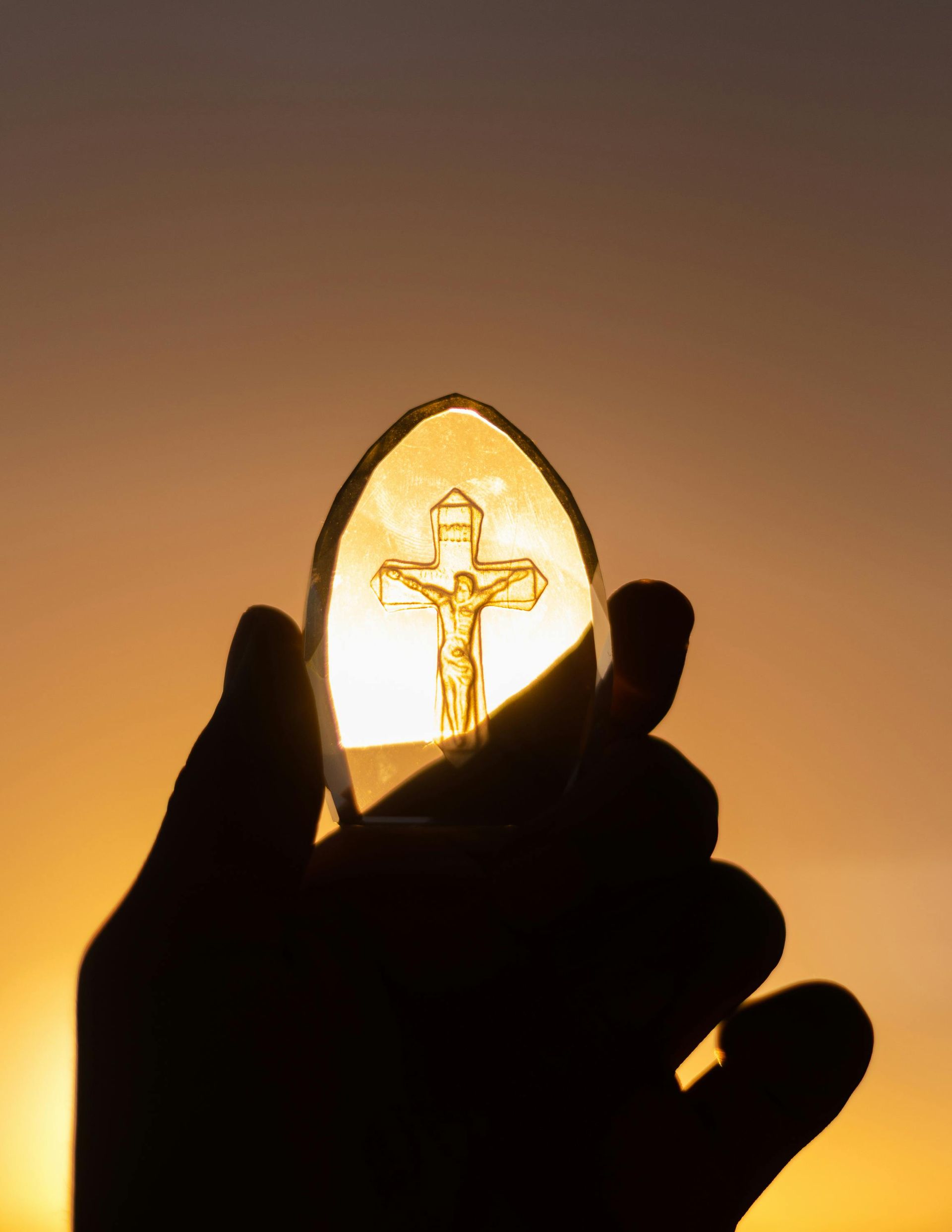 Silhouette of a hand holding a glowing crucifix against a sunset backdrop.