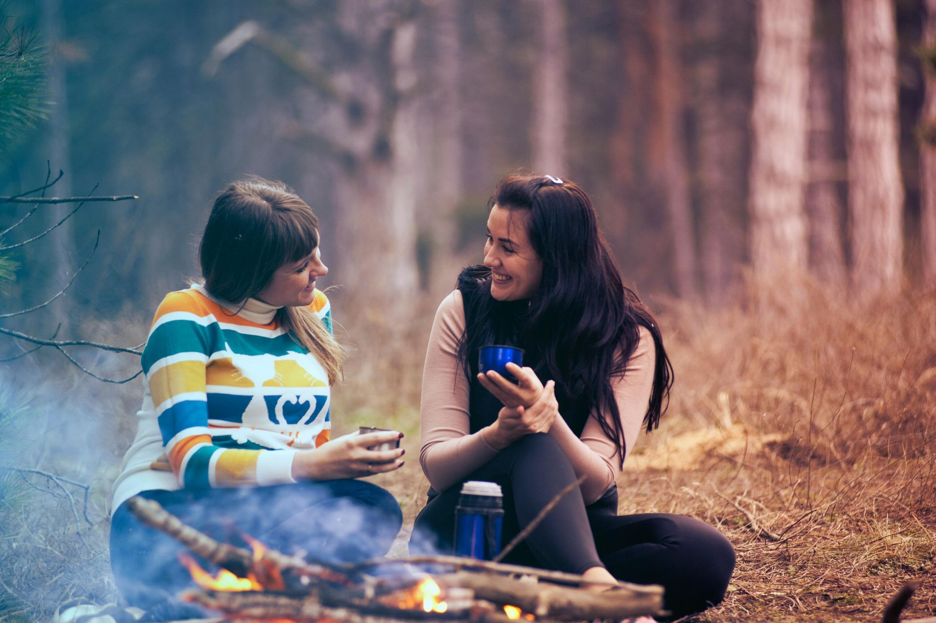 Two people sit by a campfire in a forest, conversing and holding mugs.