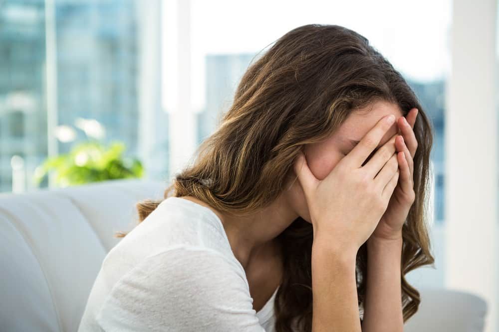 A woman is sitting on a couch covering her face with her hands.