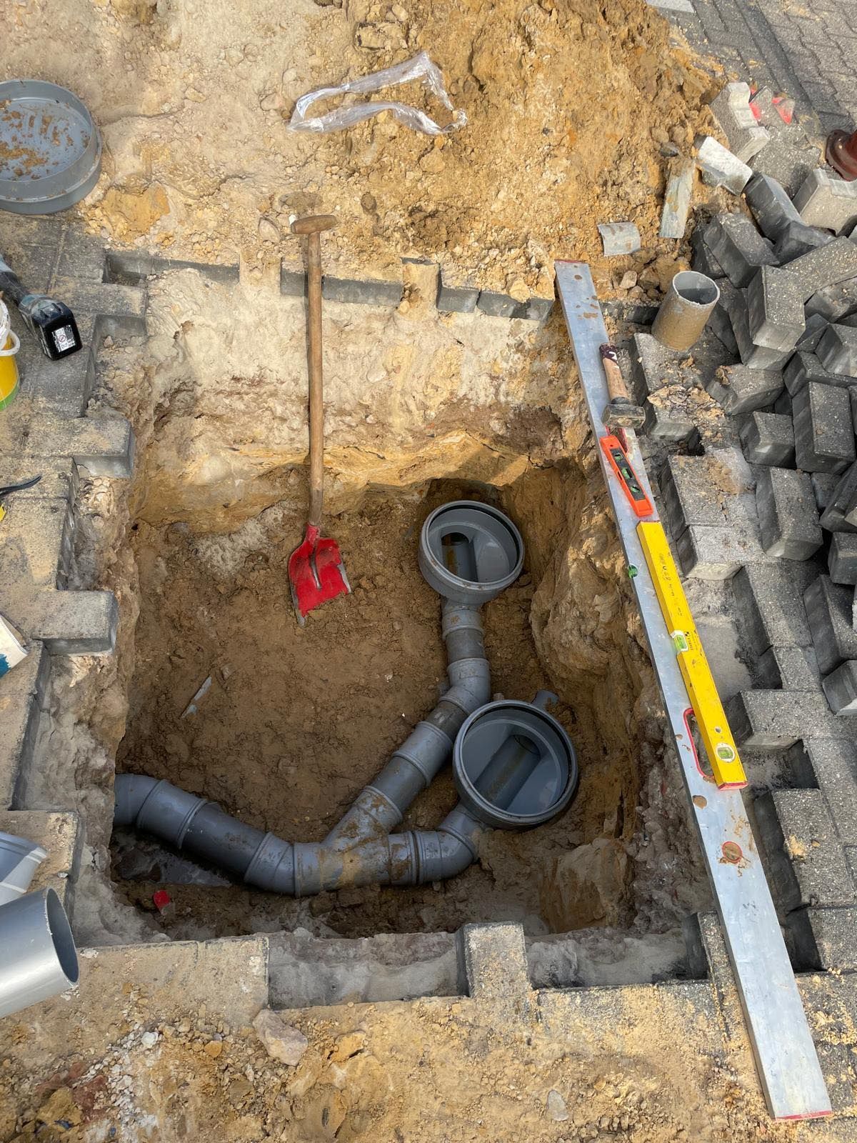 Een open sleuf met pvc-buizen en -fittingen, een schop en gereedschap in de grond naast een stenen muur.