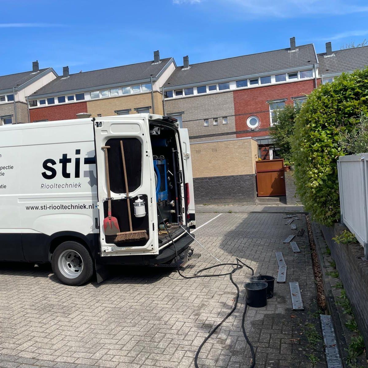 Witte bestelbus met open achterdeuren in een woonstraat naast bakstenen huizen en bestrating.