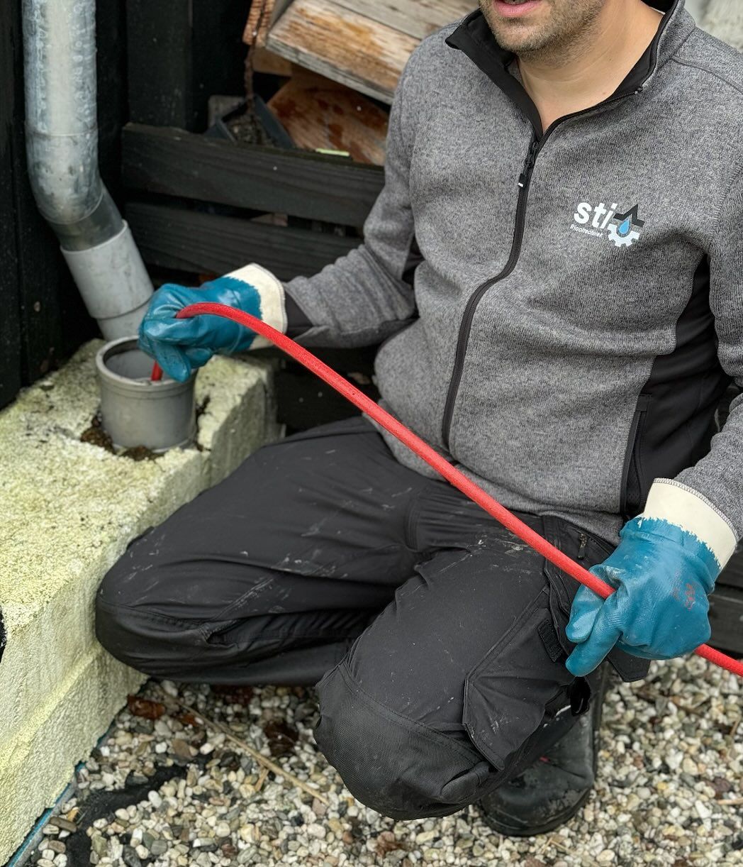 Een arbeider knielt bij een afvoerput, met een rode staaf en blauwe handschoenen naast een grijze buis.