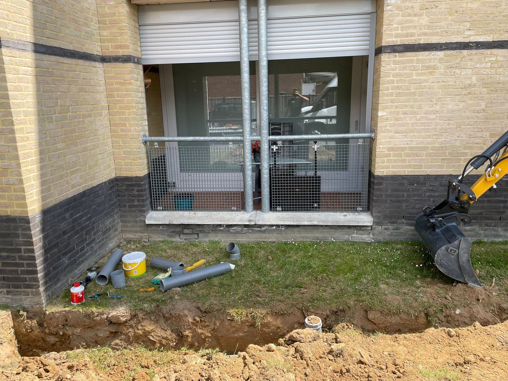 Een sleuf in de bouwput langs de muur van een gebouw, met gereedschap en een graafbak naast een raam.