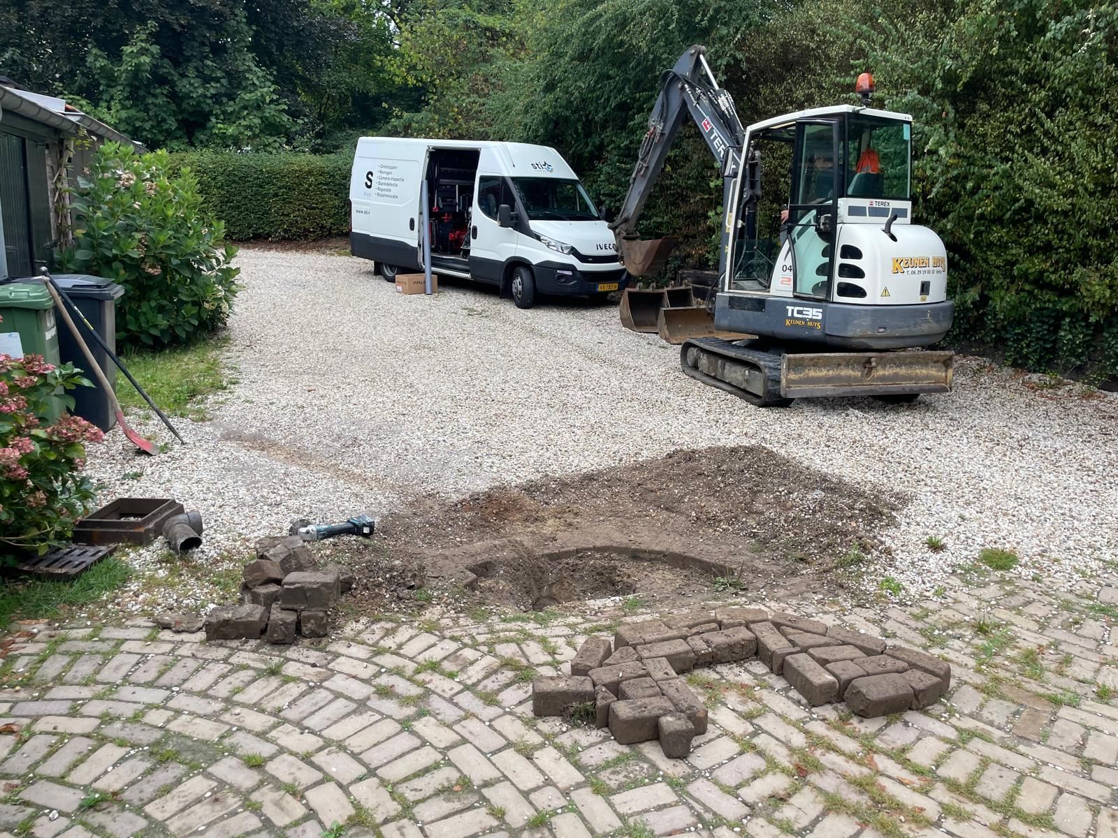 Witte bestelwagen en kleine graafmachine op een grindpad met straatstenen op de voorgrond.