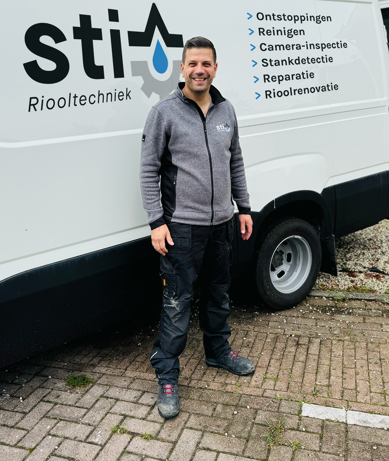 Een man staat naast een witte servicebus met Nederlandse tekst 'reparatie/service', buiten op een stoeptegel.