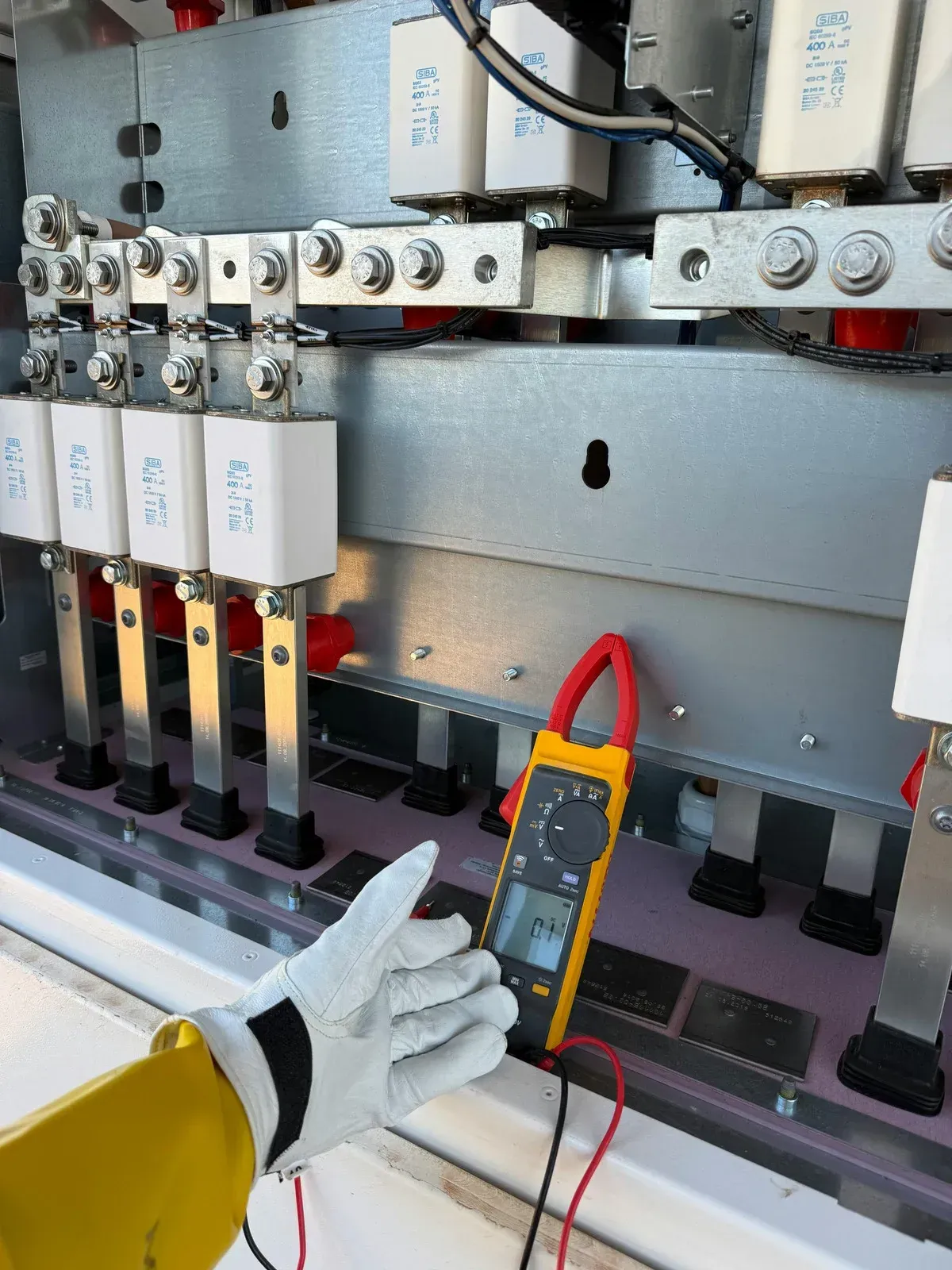 A gloved hand uses a multimeter to measure electrical components in a panel.