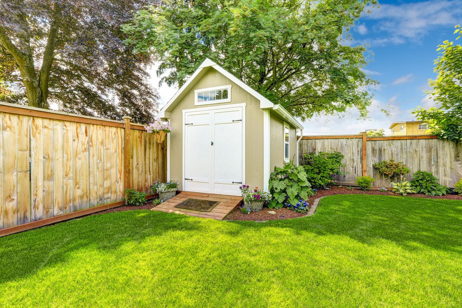 A small shed with white doors and a window in a fenced-in, grassy backyard.