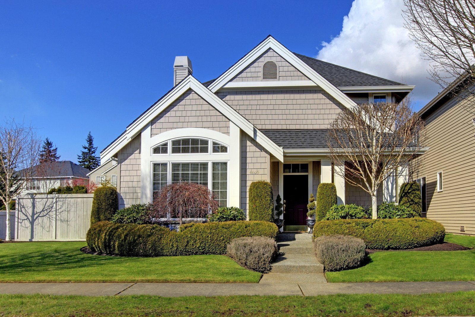 House with gray siding, arched window, and manicured landscaping under a blue sky.